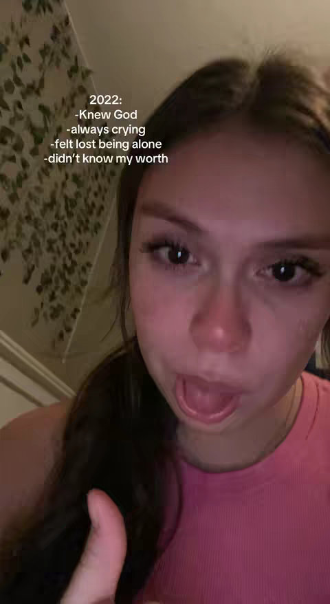 A selfie-style shot of a young woman, seen from the chest up. She has long, dark hair, and is wearing a pink ribbed top. Her face shows evidence of crying, with red around her nose and eyes. The background is slightly out of focus, showing a wall with what appears to be leafy decorative elements. Text is overlaid on the image, listing various emotional states.