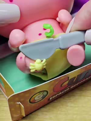 The cover image shows a close-up of a toy, possibly a Nerlie Baby, being examined. A hand holds a dull gray knife over its abdomen, above a small yellow crab-like accessory. The toy is pink and has visible belly. The toy sits in a partially visible cardboard box labeled with text in a stylized font. The overall composition highlights the toy and the interaction with it.