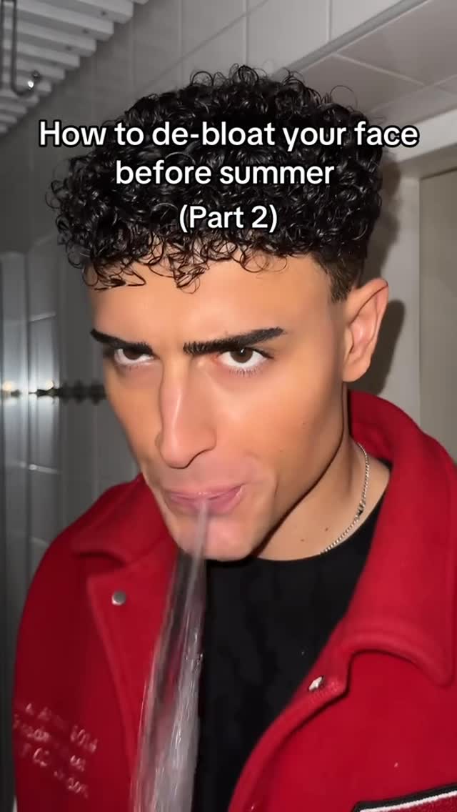 The cover image shows a young man, likely the content creator, in a close-up shot. He is positioned close to the camera, with his face taking up most of the frame. He has dark, curly hair that is neatly styled. The man is wearing a red jacket over a black shirt and a silver necklace. The background is blurred, showing what appears to be a white tiled wall. A stream of water is coming out of his mouth, indicating he is spitting water. Text overlayed at the top of the image reads, How to de-bloat your face before summer (Part 2).