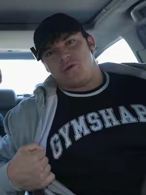 A man in a black baseball cap and Gymshark t-shirt is seated inside a car. The interior of the car and light from outside are visible. He appears to be looking directly at the camera with a slightly open mouth.