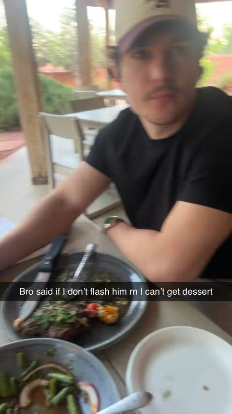 A man wearing a baseball cap and a black shirt is seated at a table, appearing to be at an outdoor restaurant. Plates of food, including a meal of meat and vegetables, are visible in the foreground. The background is slightly blurred, suggesting a casual setting, with glimpses of greenery and outdoor seating.