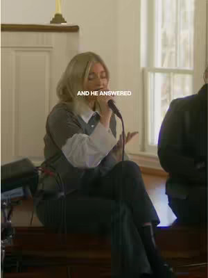 The image features a woman singing with a white shirt and gray jacket, holding a microphone in a church setting. Text "AND HE ANSWERED" is overlaid at the center. The background includes a window and altar.