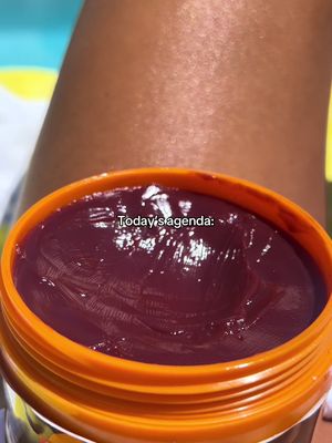Close-up shot featuring an open jar of purple cosmetic product, likely a tanning product, against a background of tanned skin. An overlay displays the text 'Today's agenda:'.