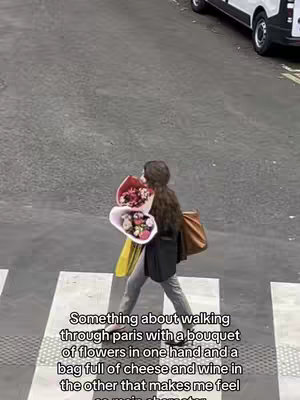 A woman walking on a crosswalk in Paris, holding two bouquets of flowers and a brown purse. The image is shot from a high angle. The woman is wearing a black top, grey pants, and a brown purse. The crosswalk is white stripes on an asphalt road, with a white van visible in the background.