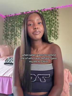 A woman with long, dark hair poses in front of a bedroom setting. She wears a dark blue tube top with white text. Text across the bottom says, 'when satan wants to remind me of who I was before I gave my life to Christ'. Behind her is a bed and a wall decorated with greenery.