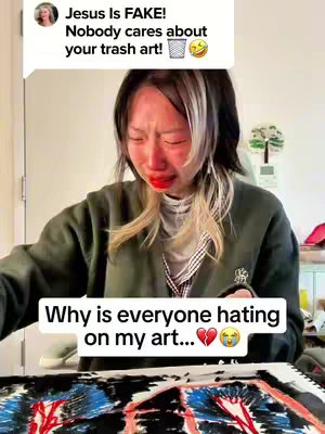 The image shows a young woman with medium-length black hair with blonde highlights, crying and is sitting in front of a drawing. She is wearing a green cardigan over a white shirt. Above her, a speech bubble shows a negative comment about her art.