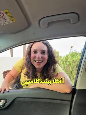 A young woman leans out of the open window of a car, smiling directly at the camera. The car interior is visible, with the dashboard and ceiling. A text overlay in Arabic appears on the image. The background contains greenery.