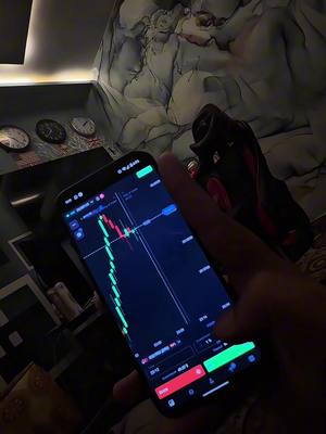 A person is holding a smartphone displaying a trading app interface. The screen shows red and green lines, a chart with data points, and control buttons at the bottom. The phone is in portrait orientation. The background is dimly lit, with a blurred effect. There is some pattern on the wall in the background. The phone is being held by a hand where the color is relatively dark. The scene gives a sense of depth through the blurred background