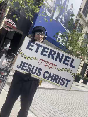 A man wearing a black cap and glasses holds a sign in a city setting. The sign displays the word "ETERNEL" at the top, a line of Hebrew script in the middle , "JESUS CHRIST" at the bottom, with DNA inspired details on the sides.