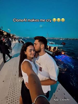 A couple is embracing on a waterfront at dusk. The man is kissing the woman on the head. The background includes a body of water with boats, city lights in the distance, and a dark blue sky.