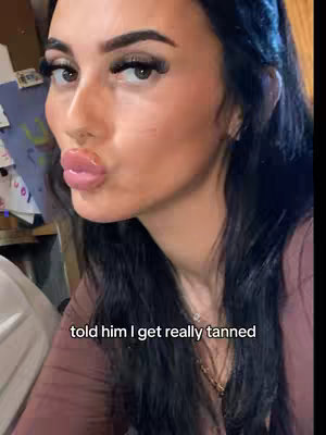 A selfie-style shot featuring a young woman gazing slightly off-center with a playful expression. Her lips are noticeably glossy and pursed, possibly in a kiss gesture. Her dark hair cascades down her shoulders, and the background reveals a glimpse of an interior setting with a cluttered workspace or storage area.