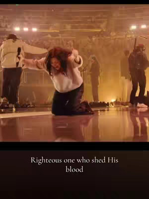 A woman kneels on a stage, hair flowing, arms outstretched. Other people are in the background, blurred, with warm stage lighting.