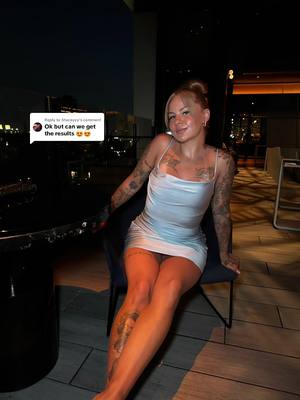 A young woman with blonde hair, in a bun, is sitting on a chair on a terrace at night. She is wearing a short, light blue dress and has tattoos on her arms and legs. The background shows city lights.