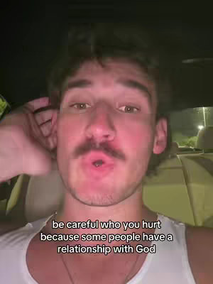 A selfie-style shot with a man in a white tank top inside a car. The man has short, dark hair, a mustache, and a slight smile. The background suggests the car's interior.