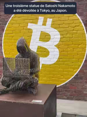 A photo shows a black metal sculpture of a hooded figure seemingly using a laptop sitting on a brown rectangular block. The backdrop is a brick wall with a large yellow circle painted on it, featuring a white Bitcoin symbol. Above the sculpture, the text announces the unveiling of a Satoshi Nakamoto statue in Tokyo, Japan.
