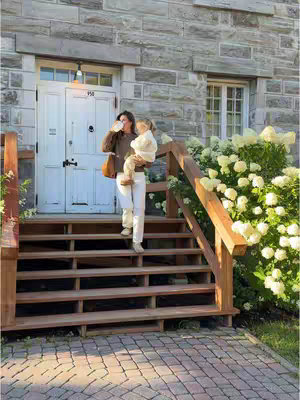 A well-lit, natural outdoor shot captures a woman descending wooden stairs, holding and carrying a child, with a background of a stone building, greenery, and bright sunlight. The woman, center-frame, wears a dark brown sweater, white pants, and a brown shoulder bag, while the child is wrapped in a cream-colored jacket and is seated in her arm . The stairs and the side of a building are in view.