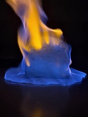 A square of bread is engulfed in flames. Blue flames emanate from the bottom, with streaks of yellow and orange reaching upwards. The fire is reflected on a glossy, dark surface.