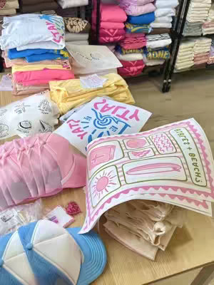 The image shows a wooden table covered with various t-shirts, hats, and printed sheets of designs, likely for a retail display. The items are mainly light-colored, with pink and blue accents. There are printed papers and designs. The items are positioned to show off retail products. There's a hint of shelving in the background, suggesting a store setting.