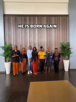 A group of seven people, three men and four women, are standing in a line. They are dressed in both formal and semi-formal attire. The background is a wall with vertical wooden paneling. Text overlayed near the top reads "HE IS BORN AGAIN."