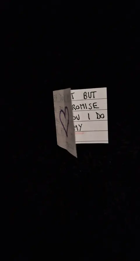 A close-up shot against a dark background features a slightly crumpled, partially folded note. The note is white with handwritten text visible. A large, stylized heart is drawn in the center, and the text is written in black ink. The angle suggests it might be a secret note.