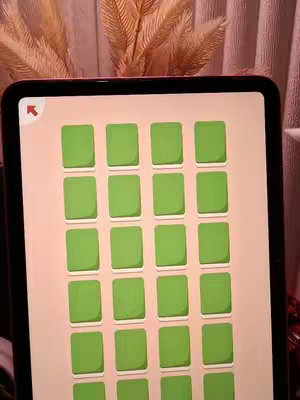 An iPad screen displaying a puzzle game in a vertical orientation. The background is a light peach color upon which green rectangular tiles are evenly arranged. A soft background of decorative leaves and soft lighting are behind the iPad.