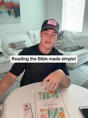 A man sits at a round table with a bible open in front of him. He is wearing a black cap and t-shirt. The text overlay on the video says 'Reading the Bible made simple!' The background includes a white couch and a window.