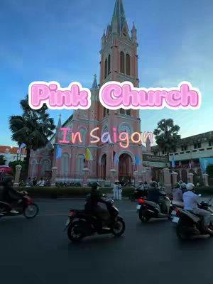 A photograph of a pink church in Saigon, Vietnam. The church is a tall, Gothic-style building with multiple spires and windows. In the foreground, there are several motorcycles on the road. The sky is a clear blue, providing a striking contrast to the pink building.