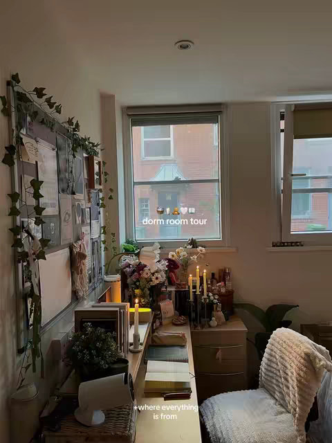 A well-lit dorm room interior, viewed from a low angle. A window on the right reveals a glimpse of an exterior view. Against the left wall is a collage of photos and notes behind a decorative vine. A desk with books and candles is positioned beneath the window, with a chair in frame right. The room is warm-toned with soft lighting and a cozy aesthetic.