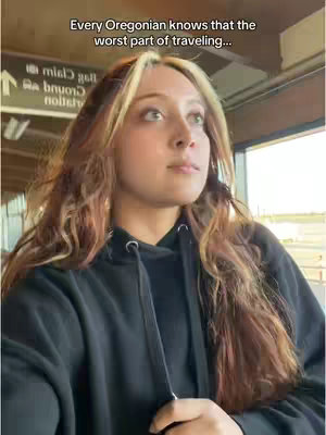 A young woman with long, wavy blonde-highlighted brown hair looks upwards, appearing thoughtful or contemplative, in what looks like an airport or train station. She wears a black hoodie.