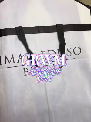 A close-up shot of a white garment bag with black handles and straps. The bag has text on it, and a multi-colored text overlay with 'GRWM AMVCA11 Part 1' is placed over it. The bag appears to be made of a smooth, possibly satin or similar material.