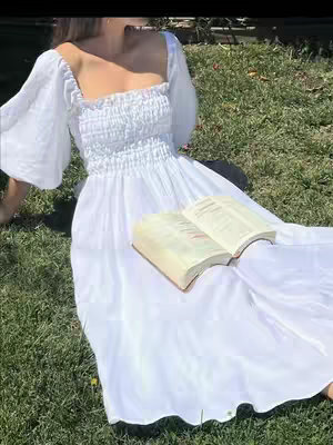 A woman in a white dress with puffy sleeves and a square neckline is sitting in a grassy area. She is holding an open book in her lap. The dress drapes around her on the grass. The image is well-lit and warm in tone.