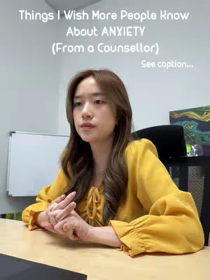 A woman with long brown hair is seated at a table with her hands clasped in front of her. She is wearing a yellow blouse. Behind her is a white board and a piece of art.