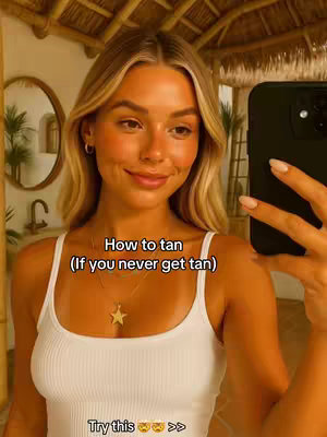 A selfie-style shot of a fair-skinned young woman in a white tank top, indoors. She is holding a black phone, angled towards her face, and shows a slight smile. The background has a wooden ceiling and a round mirror on the wall.