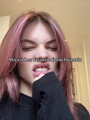 A close-up of a person with pink hair, slightly angled, with text overlayed. The person is looking towards the side with a subtle smirk, their mouth slightly open, and fingers touching their mouth conveying a thoughtful expression. The background appears to be a neutral interior, contributing to the focus on the subject's face. The lighting seems natural, emphasizing the skin tones and the pink hair.