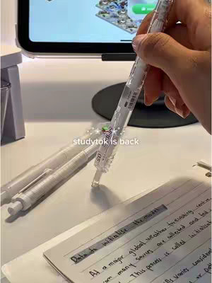 The image shows a workspace setup for studying. In the foreground, a hand is holding a clear pen over an open notebook with handwritten notes. Two other similar pens are lying to the left. Behind the pens and notebook, there's a partially visible iPad displaying a screen, and in the background, there is a black desk lamp. The overall composition is simple and clean, with a focus on study materials.