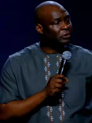 A man with a shaved head is shown from the chest up, appearing to speak with a microphone in hand. He's wearing a dark gray shirt with a detailed white floral embroidery pattern down each side of the chest.