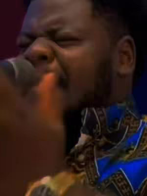 A close-up shot captures a Black man passionately singing into a microphone. He's wearing a patterned blue and gold shirt. His eyes are closed, conveying deep emotion, and his face is contorted as he sings, with his mouth open and hand close to the mic.