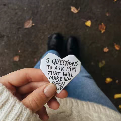 A hand holding a white paper heart with handwritten text, set against a background of legs in jeans and black shoes, and a dark ground with scattered leaves.