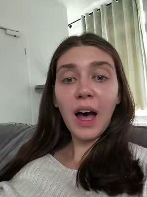 A young woman with fair skin and long brown hair is featured in a close-up shot. She is looking directly at the camera with her mouth open in a surprised or excited expression. The background includes soft, neutral tones, hinting at the interior of a room..