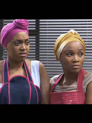 The image features two women in a medium shot, their expressions suggesting concern or contemplation. Both are looking off-camera to the right. The woman on the left wears a pink head wrap that is wrapped into a unique rose-like style, and a dark navy apron with red trim over a white top. The woman on the right has a gold head wrap, red apron and a light tan top. The background is a series of light-colored horizontal lines or blinds.