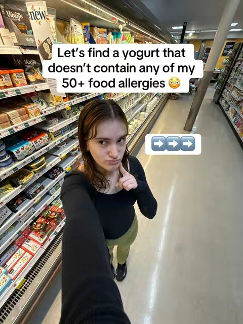 A selfie taken in what looks like a grocery store aisle. A young woman stands in the foreground holding the camera, pointing her right index finger up towards the text overlay that is placed above her head. There are shelves in the background filled with products. The colors are well-lit, and the image is composed in a way that emphasizes the woman and the text as the main focus.