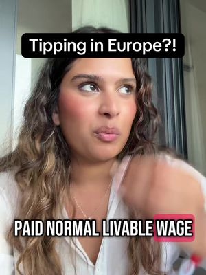 A woman with wavy brown hair looks towards the upper right, with a slightly concerned facial expression. She is wearing a white top. The image has overlaid text in a black rectangle at the top reading, "Tipping in Europe?!". At the bottom, the text "PAID NORMAL LIVABLE WAGE" is in a pink rectangular box, and there is a hand visible, performing a gesture.