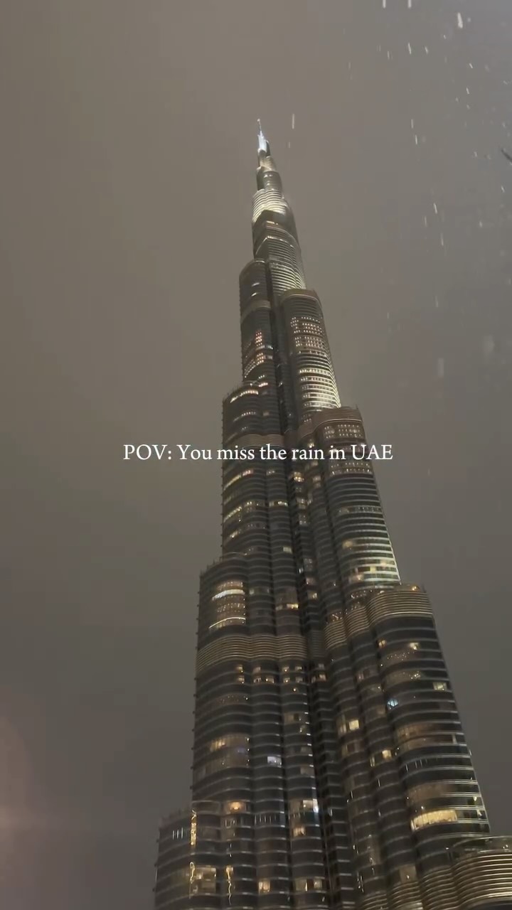 An overcast, nighttime shot of the Burj Khalifa in Dubai. The tall skyscraper is illuminated with lights, and the text overlay ‘POV: You miss the rain in UAE’ is in the center. Raindrops can be seen falling in front of the building.