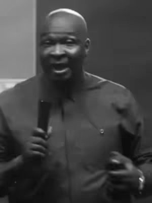 A grayscale image featuring a man speaking into a microphone. The man has short hair, and is wearing a dark collared shirt. His hands are visible with a wristwatch on one wrist is at the ready. The background is a dark wall.