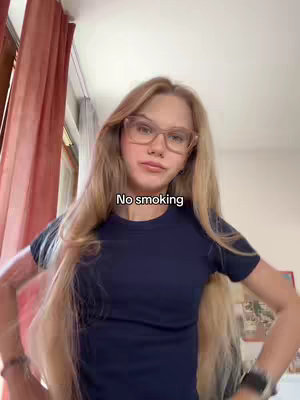 A young woman with long blonde hair and glasses looks at the camera. She wears a dark blue t-shirt and her arms are at her side. The background includes a window with red curtains.