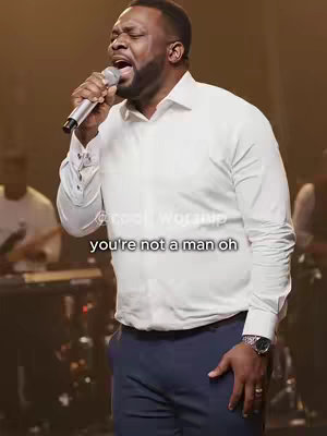 A man wearing a white dress shirt and navy pants sings into a microphone, with text overlayed on the image.