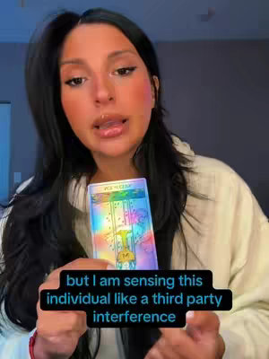 A woman with long black hair and makeup holds a tarot card, a text overlay with black background reads 'but I am sensing this individual like a third party interference.'