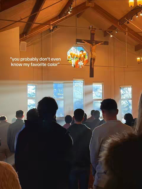 A photograph captures the interior of a church, bathed in warm, golden sunlight streaming through tall windows. The perspective is from the middle of the congregation, looking towards the altar. A multitude of people are seen from behind, their silhouettes creating a sense of depth and community. At the focal point, a large wooden crucifix hangs, with a figure of Jesus on it, illuminated by a stained-glass window and chandelier. The architecture includes wooden beams and a high ceiling, adding to the sense of reverence and light.