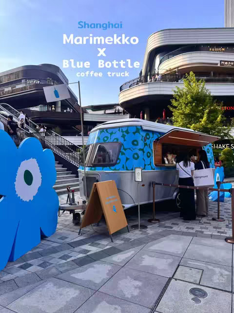 An outdoor scene with a blue and white food truck decorated with a floral pattern, parked in front of modern buildings. Text overlay in white and light blue spells "Shanghai Marimekko X Blue Bottle coffee truck" against a clear sky. People are milling about, suggesting an event or a gathering at what appears to be an upscale shopping or dining area.