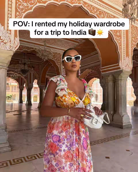 A woman in a colorful floral dress and white sunglasses is posing in a grand hall with ornate architecture. The setting is bright and sunny with warm tones.